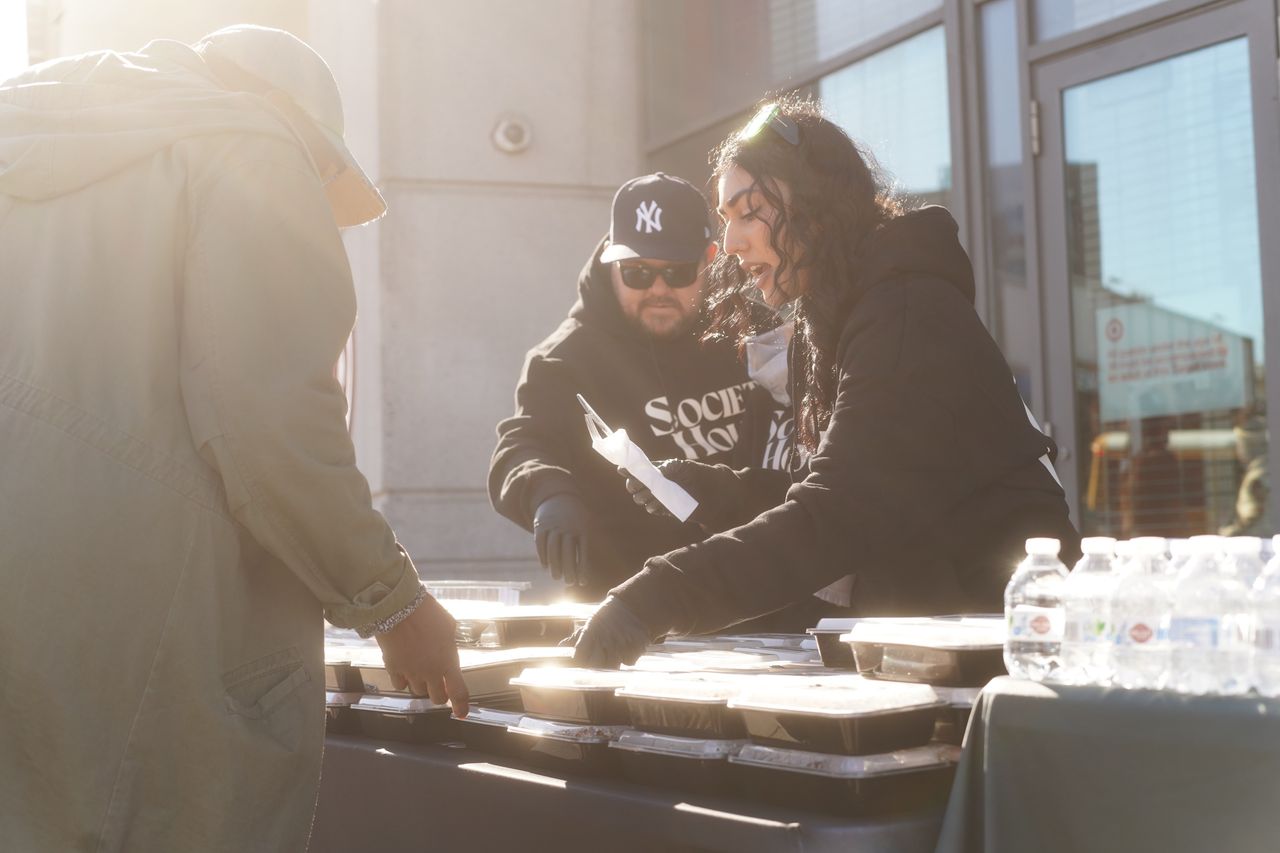 Two staff members of Society House Brooklyn standing at a table full of prepared meals in to-go containers serving someone in need of a meal.